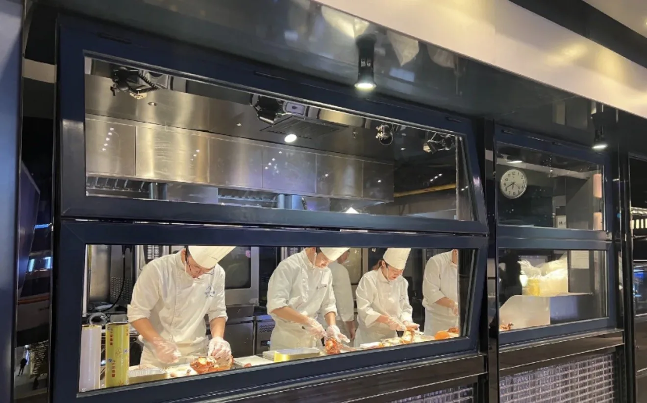A street-facing open kitchen: chefs plating behind glass with roasting units behind them, a child watching from the sidewalk